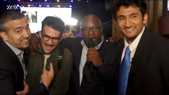 Four men posing together with smiles at an event, with a microphone held by one of them, brightly lit stage in the background.