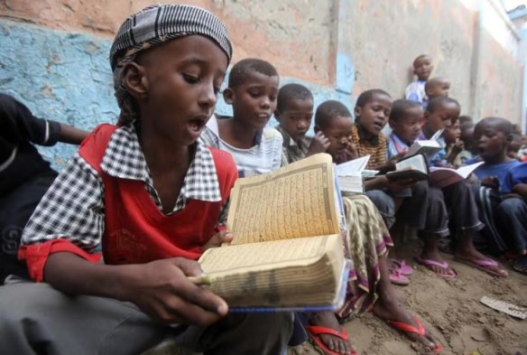 A group of children sitting in a line on the ground, focused on reading books. One child in the foreground holds an open book, reciting, while others around him are also engaged in reading.
