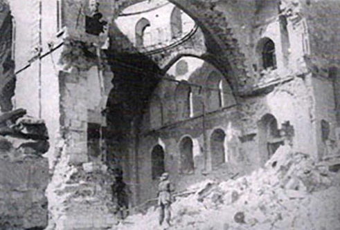 Interior view of a ruined building with crumbling walls and debris, featuring an archway and a soldier standing amidst the ruins.