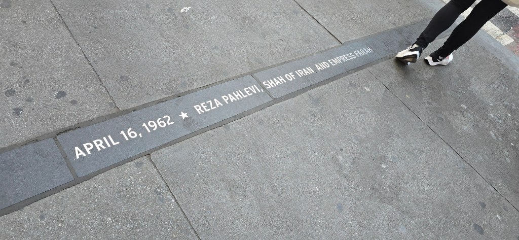 A person walking by a stone inscription on the pavement that reads 'APRIL 16, 1962 REZA PAHLEVI, SHAH OF IRAN, AND EMPRESS FARAH.'