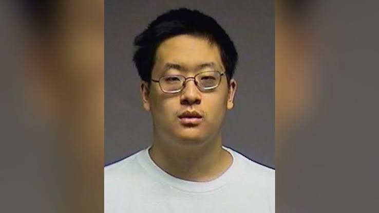 Mugshot of a young man with glasses, wearing a plain white t-shirt, looking directly at the camera.