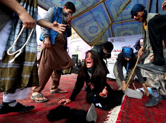 A group of people performing in a dramatic reenactment, with one individual on the ground appearing to be in distress, while others express emotions of sorrow and anger in a public setting.