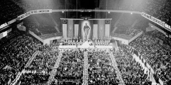 A large crowd gathered in an arena for a formal event, with a stage at the front featuring a prominent figure and decorative banners.