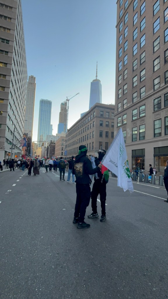 Two individuals holding a flag stand in the middle of a street surrounded by tall buildings, with a crowd in the background.