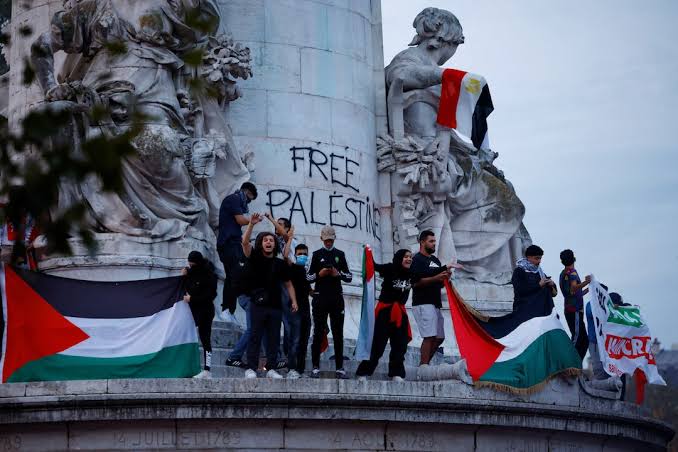 Crowd of protesters holding Palestinian flags and other banners, gathered near a monument with 'FREE PALESTINE' graffitied on it.