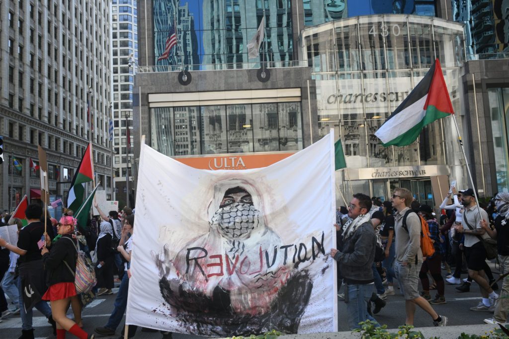 Protesters march on a city street holding a large banner that depicts a masked figure with the word 'Revolution' written on it, alongside Palestinian flags and other protest signs.