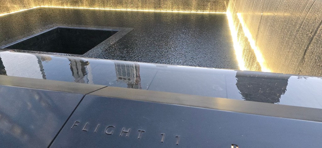 Close-up of a memorial at the 9/11 Memorial Plaza, featuring the inscription 'FLIGHT 11' on a stone edge, overlooking a water feature with a deep void surrounded by a patterned wall.